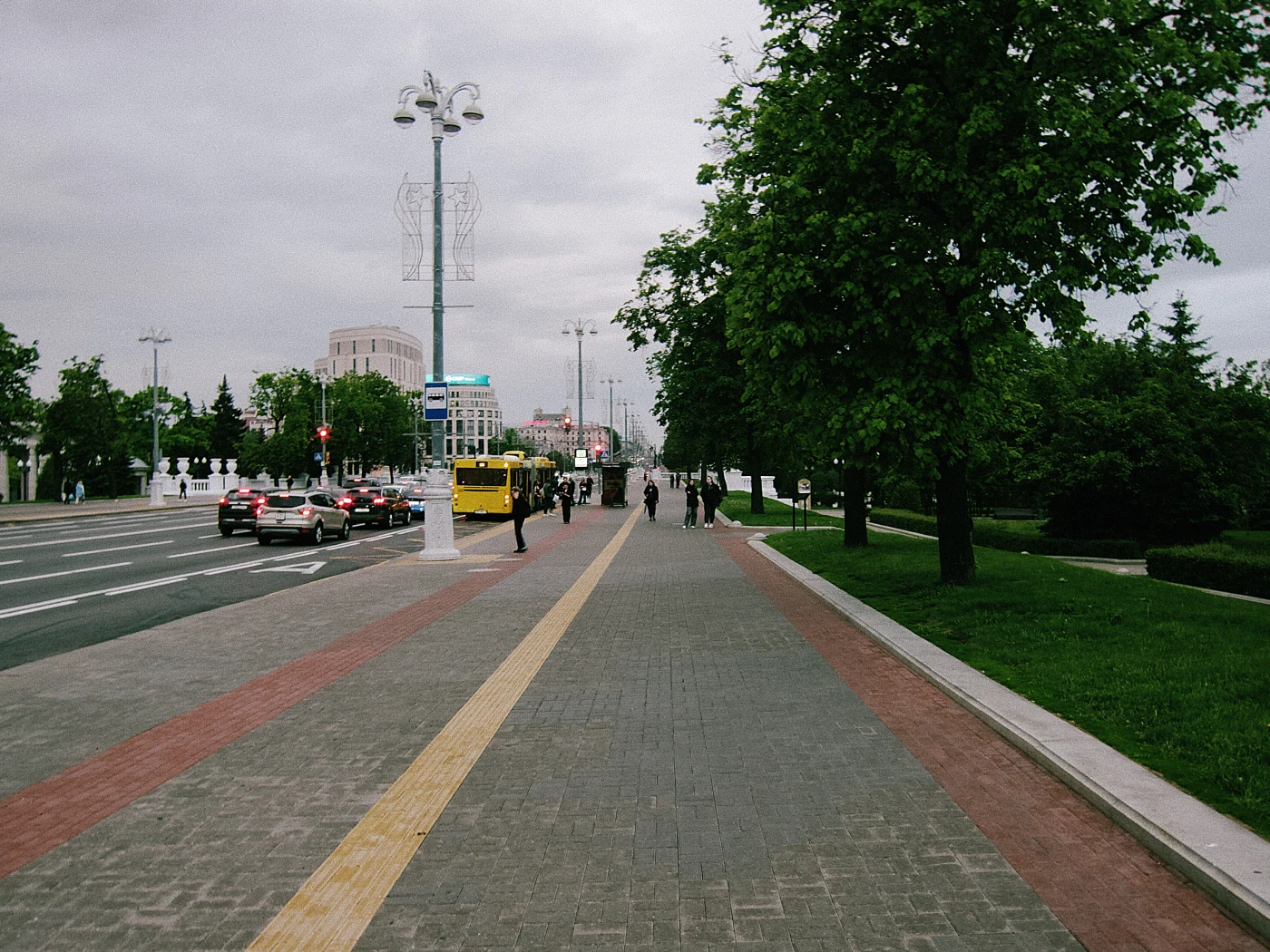 A pedestrian street with green trees, cars and people, gray sky.