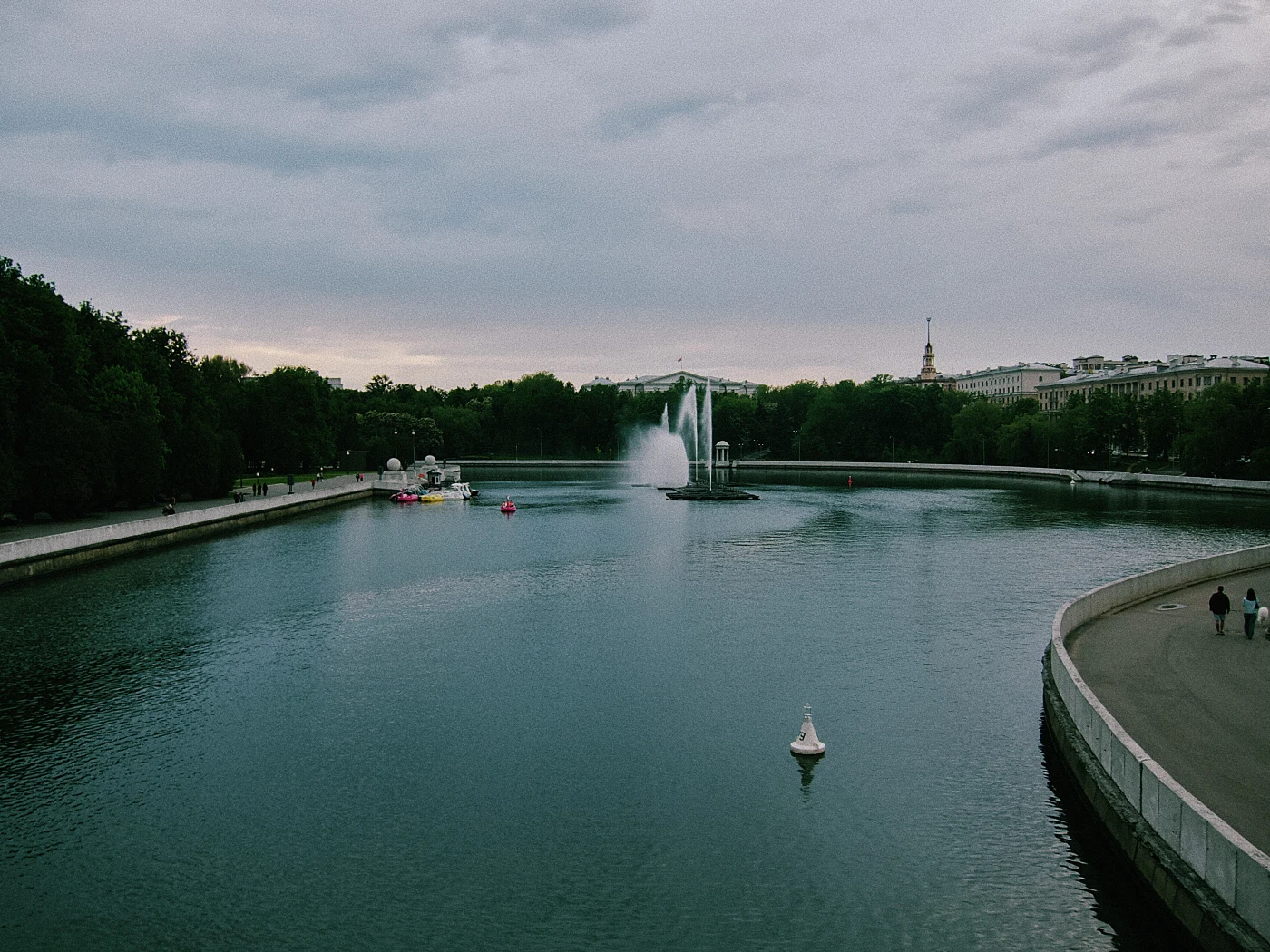 A calm pond with a fountain, surrounded by green trees and urban architecture on the horizon.
