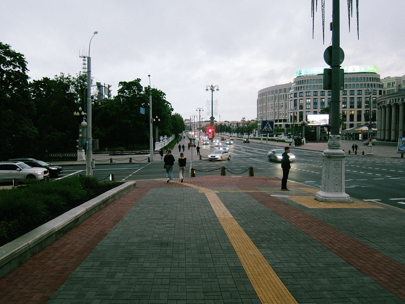 A pedestrian area with people, cars on the road, and a traffic light with a red signal. Gray sky and urban architecture.