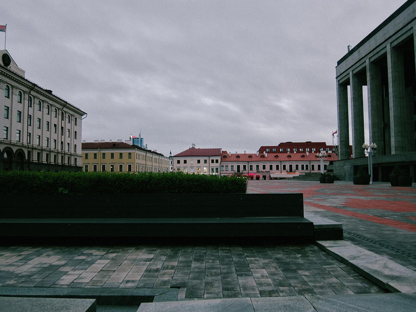 A square with a gray sky, buildings in different styles and green plantings. Columns are visible in the background.