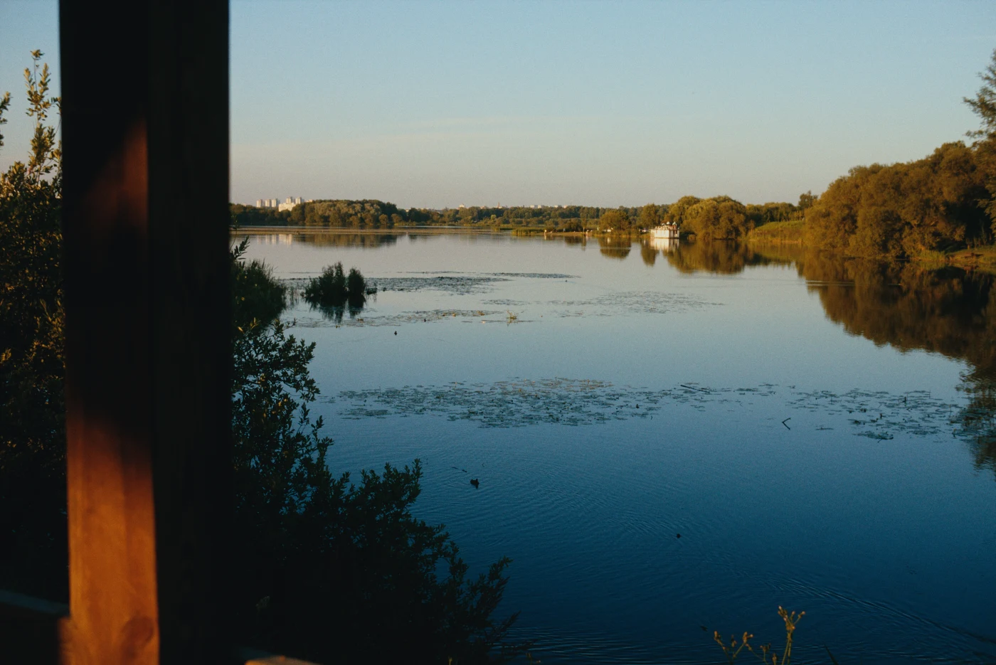 Спокойная водная гладь с отражением неба и зелеными берегами, на горизонте видны деревья и здания.