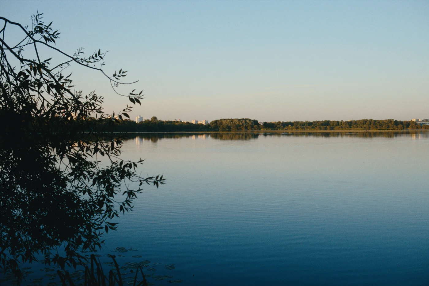 Спокойная водная гладь с отражением неба и деревьев на берегу. Вдалеке виднеются силуэты зданий.