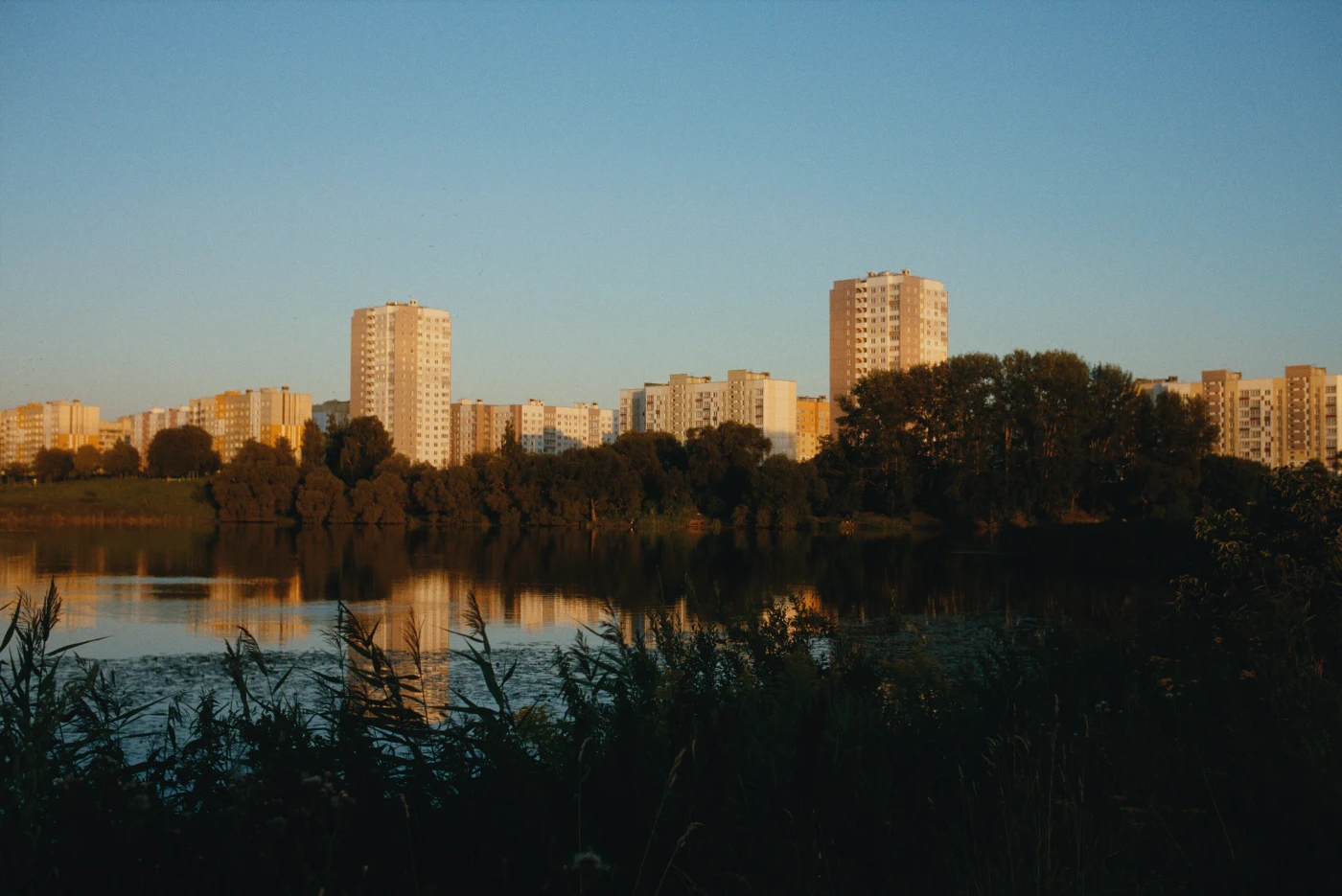 Городские высотки на фоне озера и зелени, отражение в воде, вечерний свет.