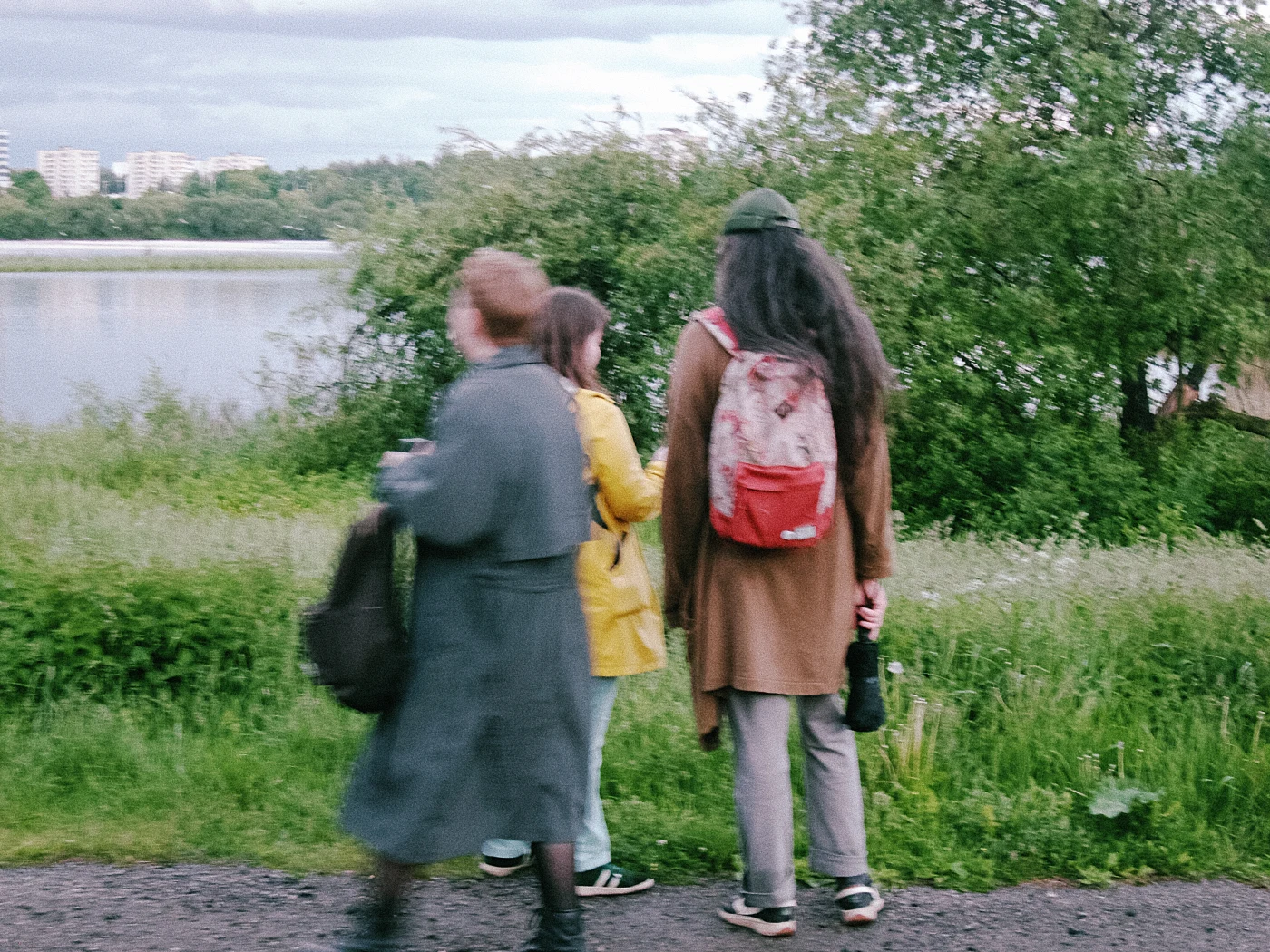 Трое людей стоят у воды, один в желтом плаще, другой с рюкзаком, третий в длинном пальто. На фоне зелень и здания.