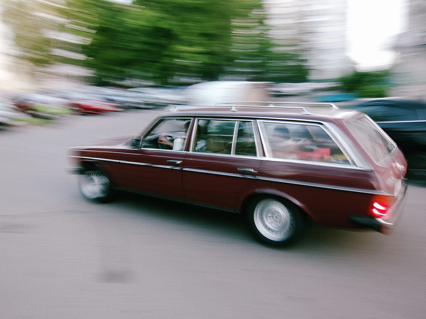 A blurred red station wagon moves down the street, with other cars and green trees around it.