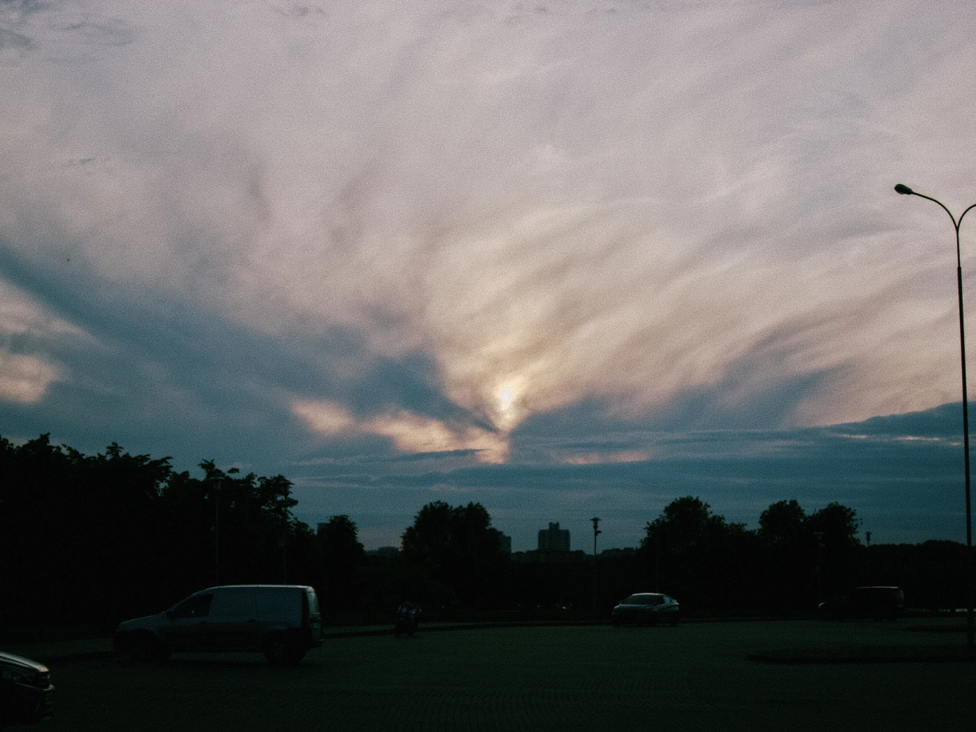Sunset with clouds, light breaking through them, with cars and trees in the foreground.