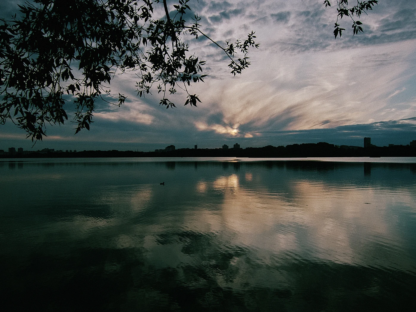 Sunset over a lake, with clouds and trees reflected in the water, creating a calm atmosphere.