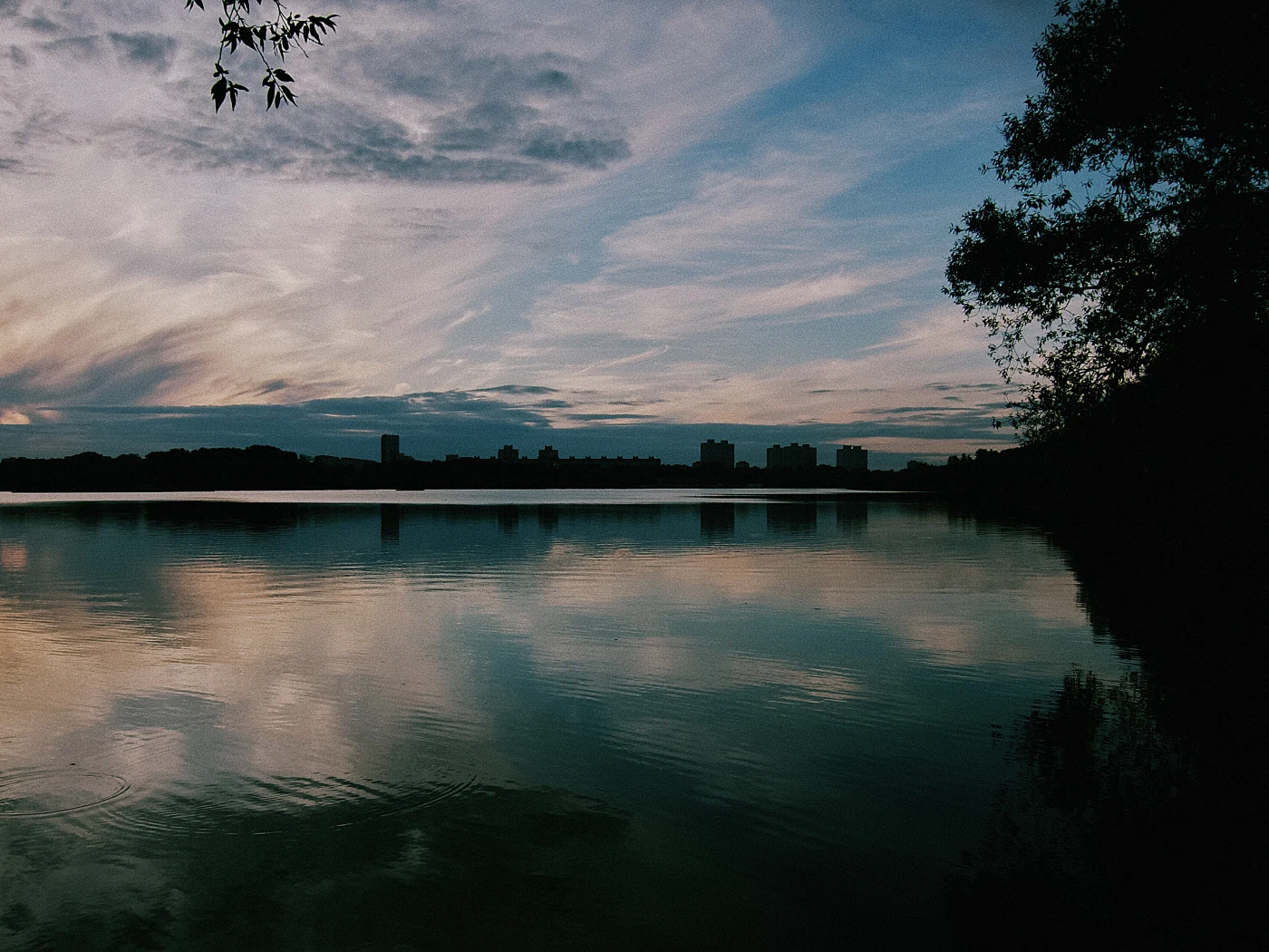 Sunset over a lake, with clouds and buildings reflected in the water, creating a calm atmosphere.