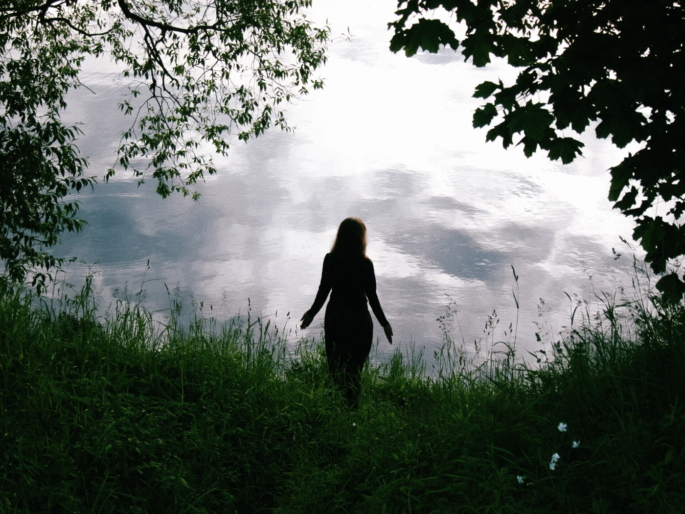 The silhouette of a person on the shore of a lake, with the sky reflected in the water and greenery around.