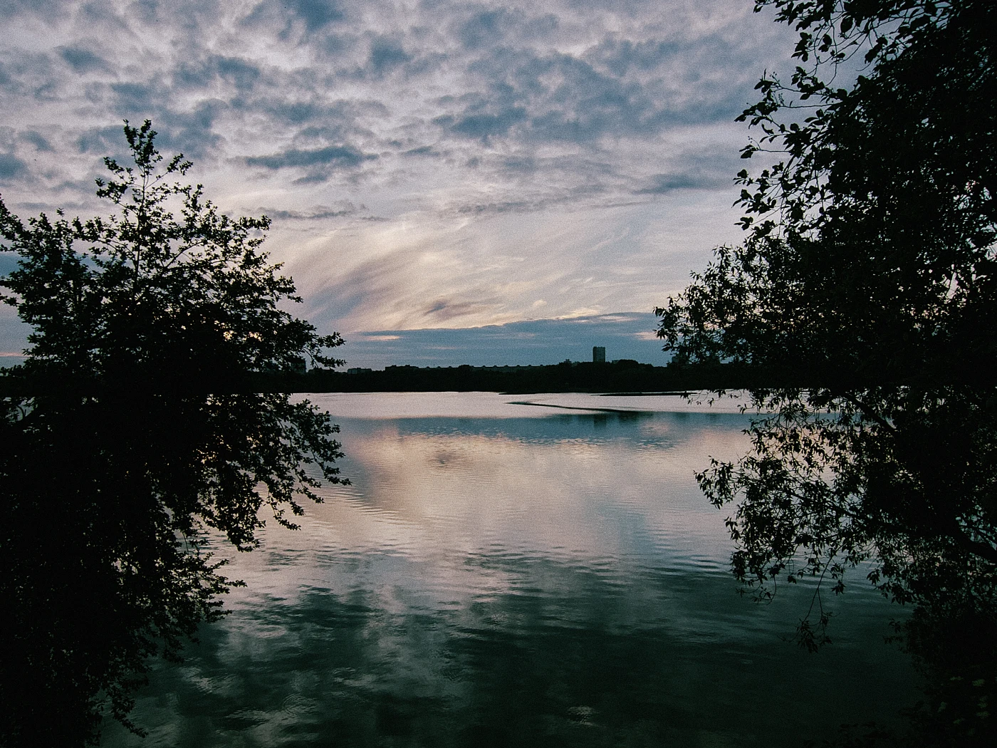 A calm lake under a cloudy sky, with trees and the horizon reflected in the water.