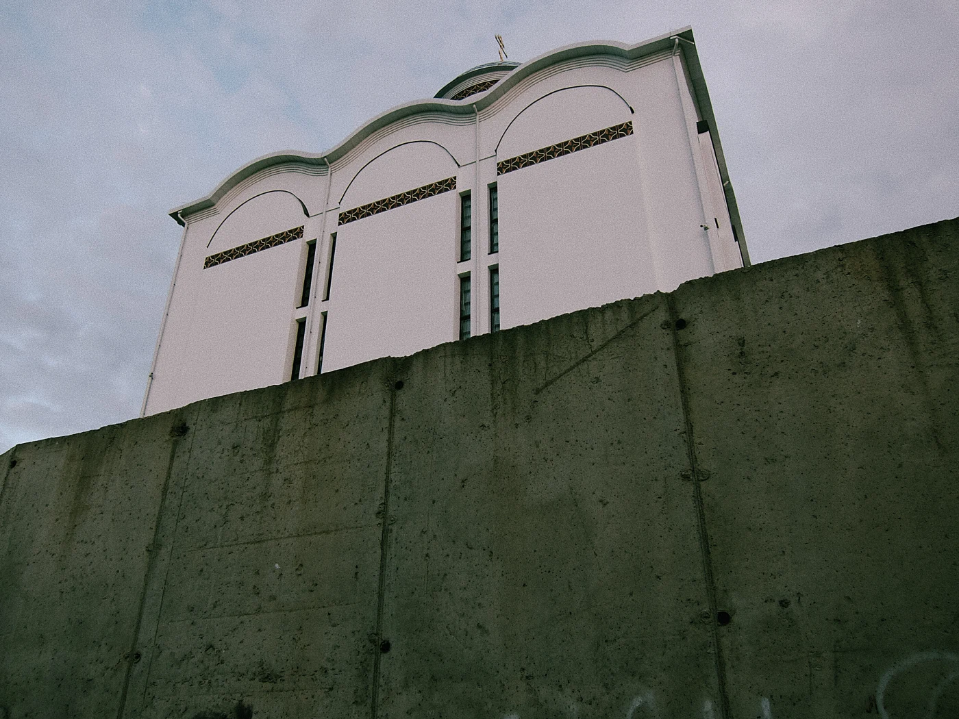 A white building with a dome, partially hidden behind a concrete wall, against a cloudy sky.
