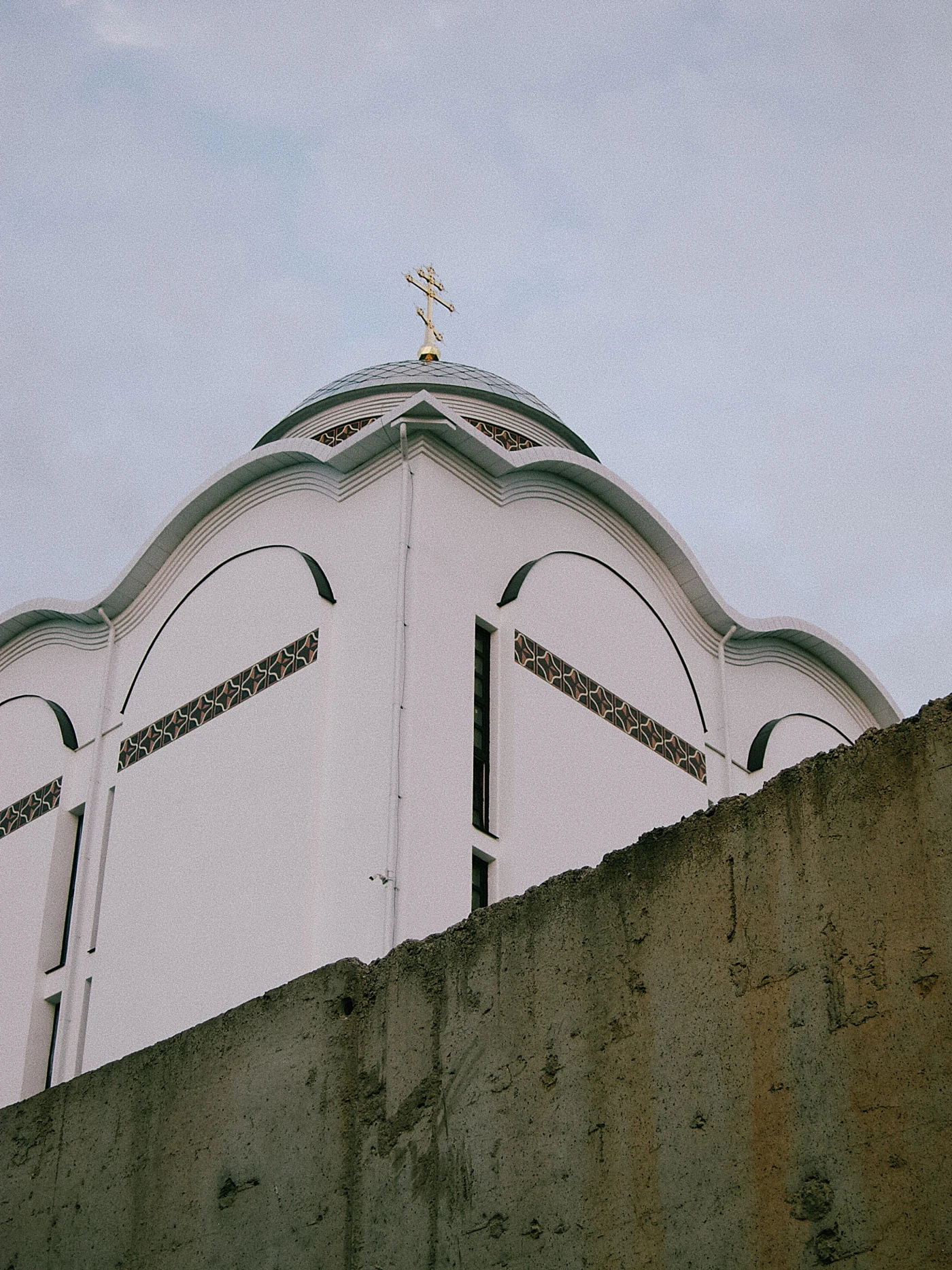 A church dome with a cross against a gray sky and a concrete wall.