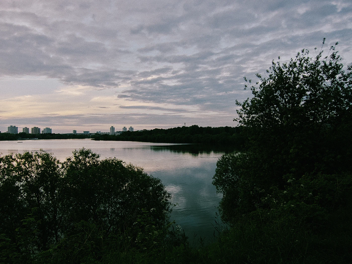 A calm lake against a cloudy sky and city silhouettes. Green trees in the foreground.