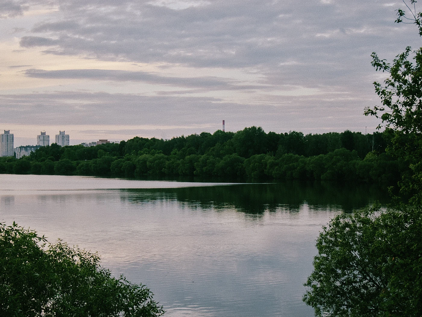 A calm river with reflections of clouds and green trees on the bank, with buildings visible in the distance.