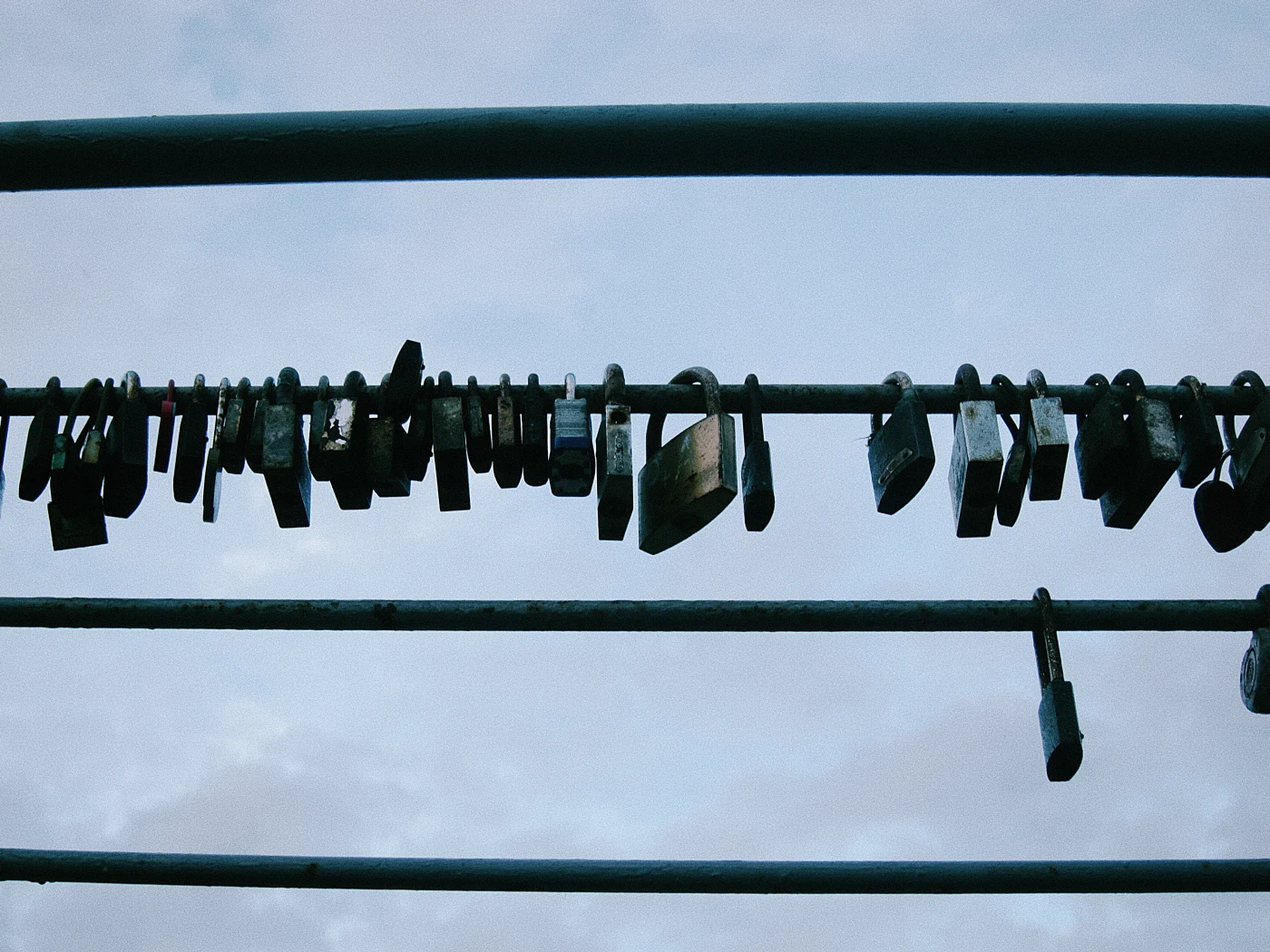 Padlocks hanging on a metal crossbar against a cloudy sky.