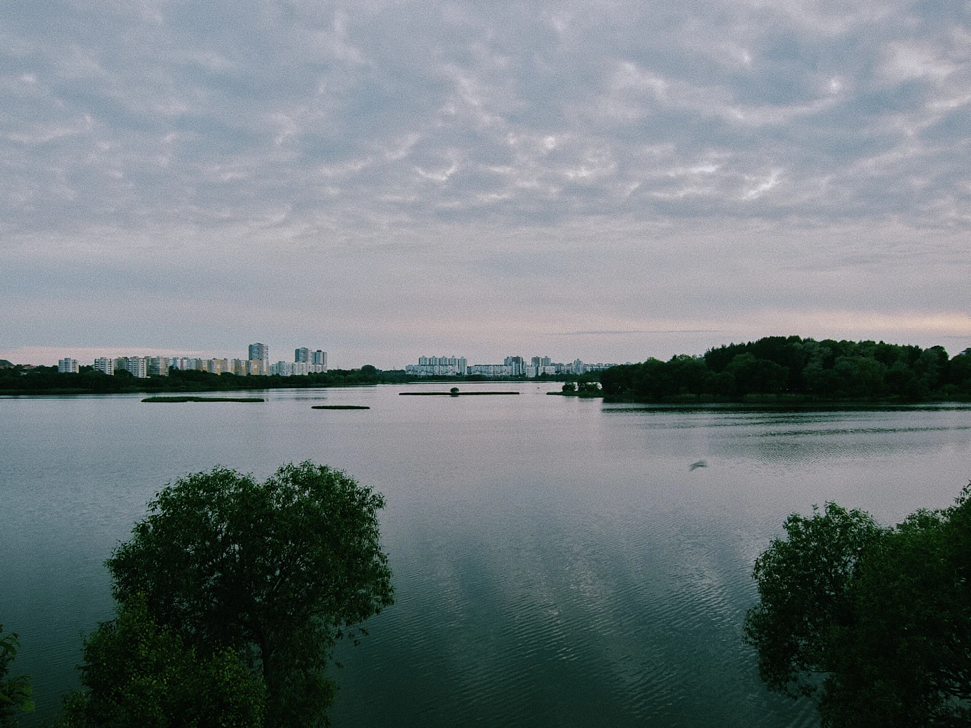 A calm river with the cloudy sky reflected in it, with city buildings and green trees in the background.