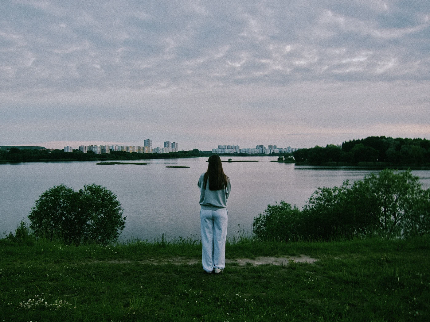 A young woman in light-colored clothing stands with her back to the water, against a cloudy sky and city buildings in the distance.