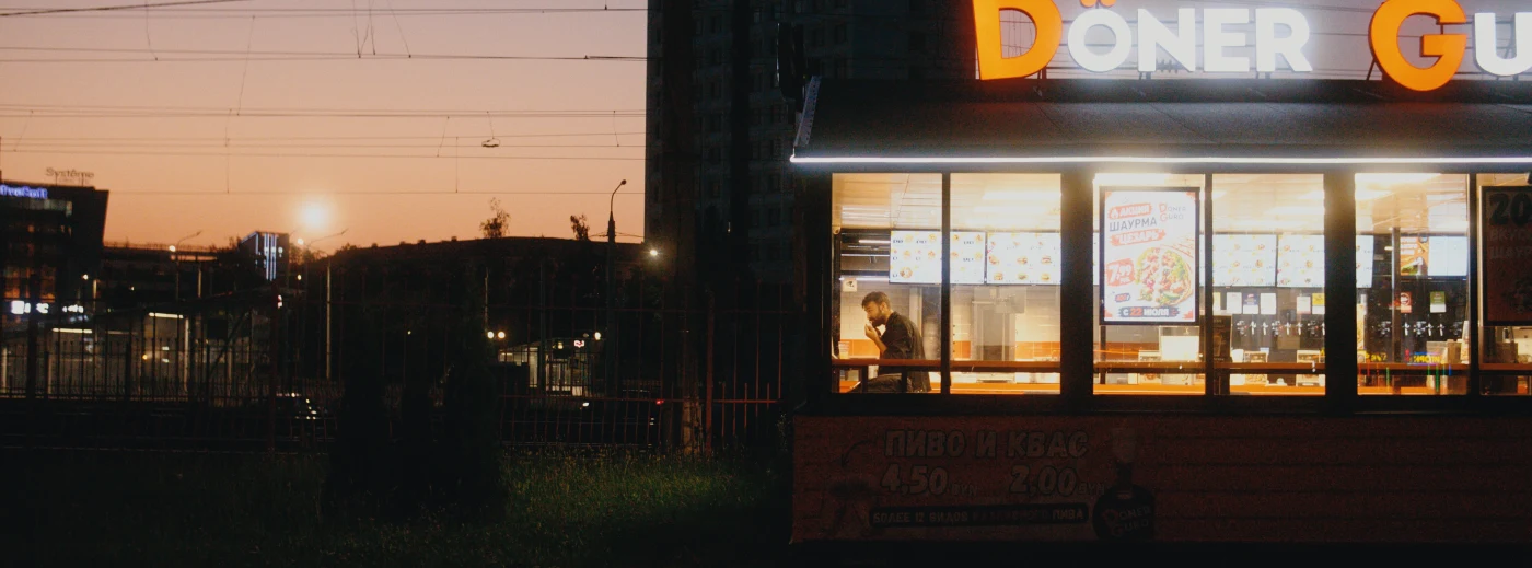 Лавка с вывеской «Döner» на фоне заката, человек внутри готовит еду.