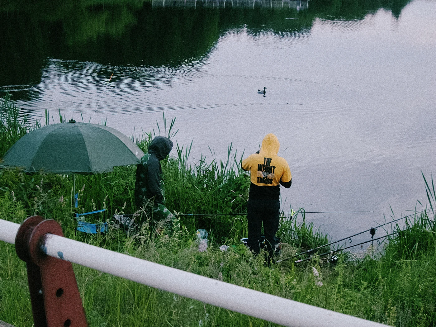 Two fishermen by the water, one in a yellow jacket and the other in green, with an umbrella and a floating duck nearby.