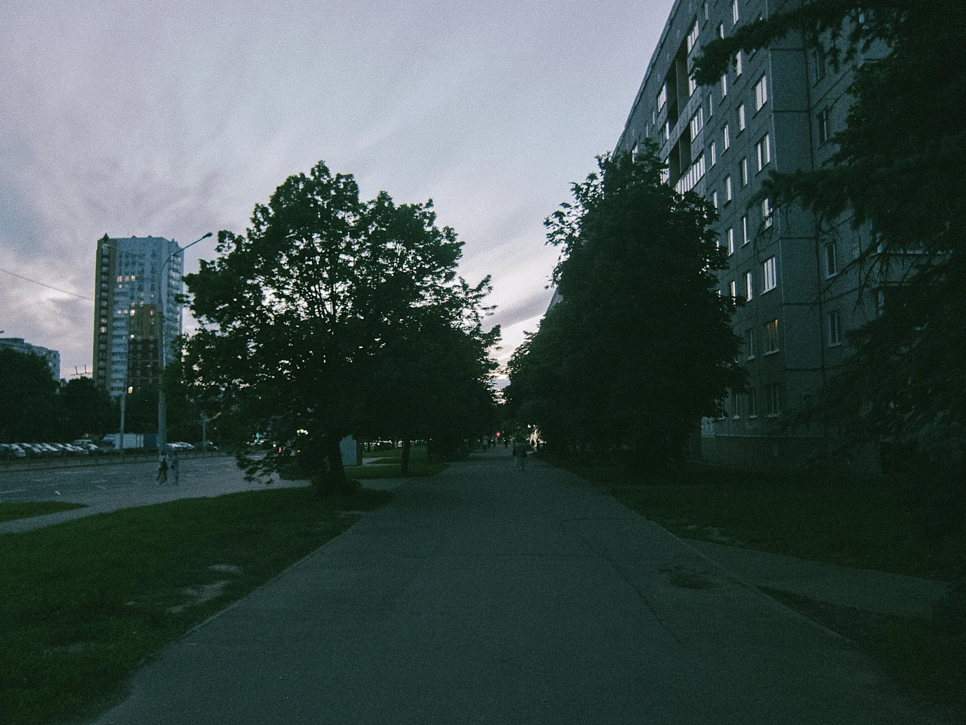 A street with trees and apartment buildings against an evening sky. People are faintly visible in the distance.