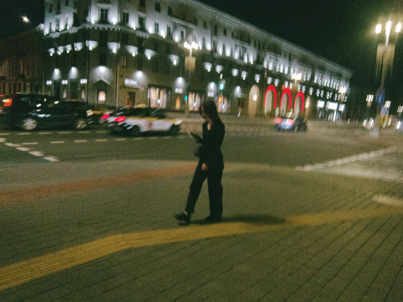 A person on a street at night, with cars moving nearby and illuminated buildings in the background.