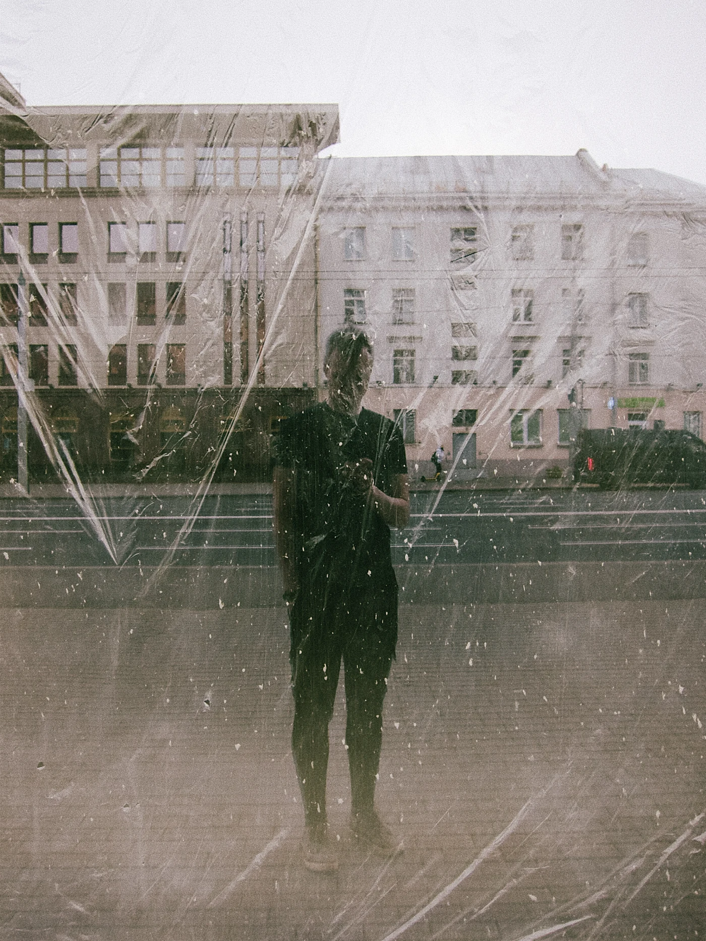 A person in black clothing stands in front of a building, reflected in fogged glass with film on it.