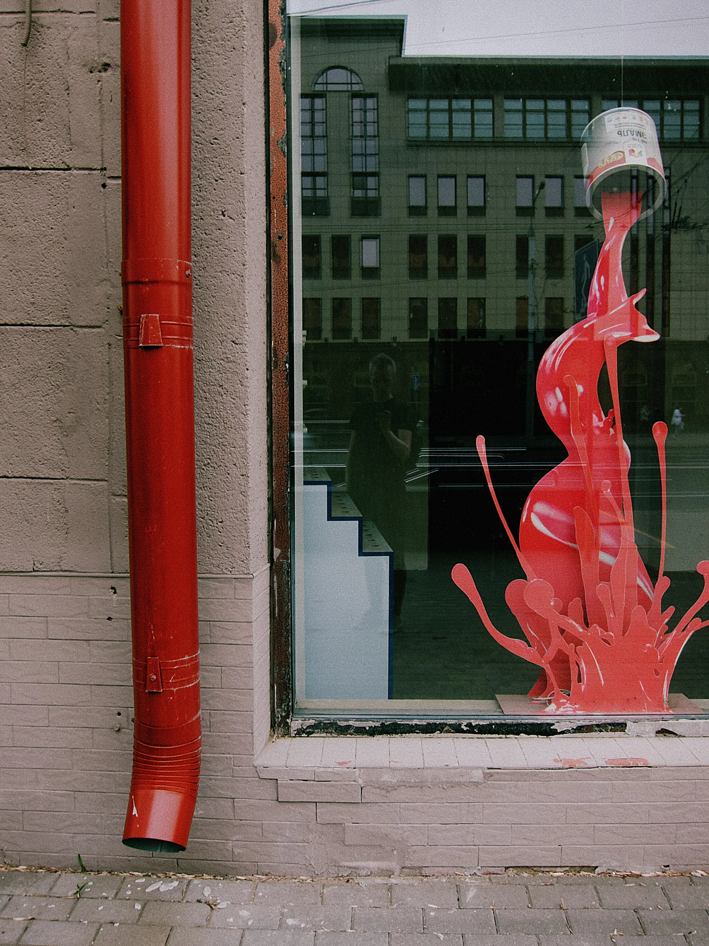 A red shop window with a figure resembling drops of paint and a building reflected in the glass.
