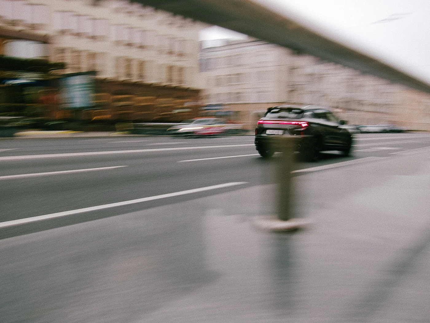 A blurred image of a black car moving along a street with road markings and a barrier.