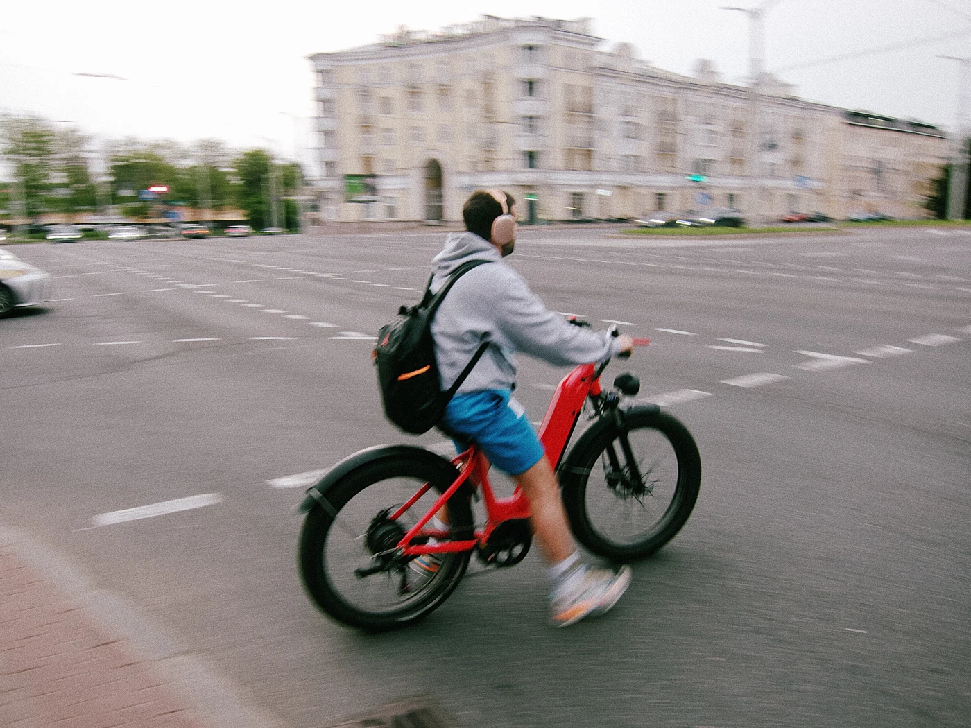 A guy on an electric bike rides through an intersection, wearing headphones and sportswear. Buildings are visible in the distance.
