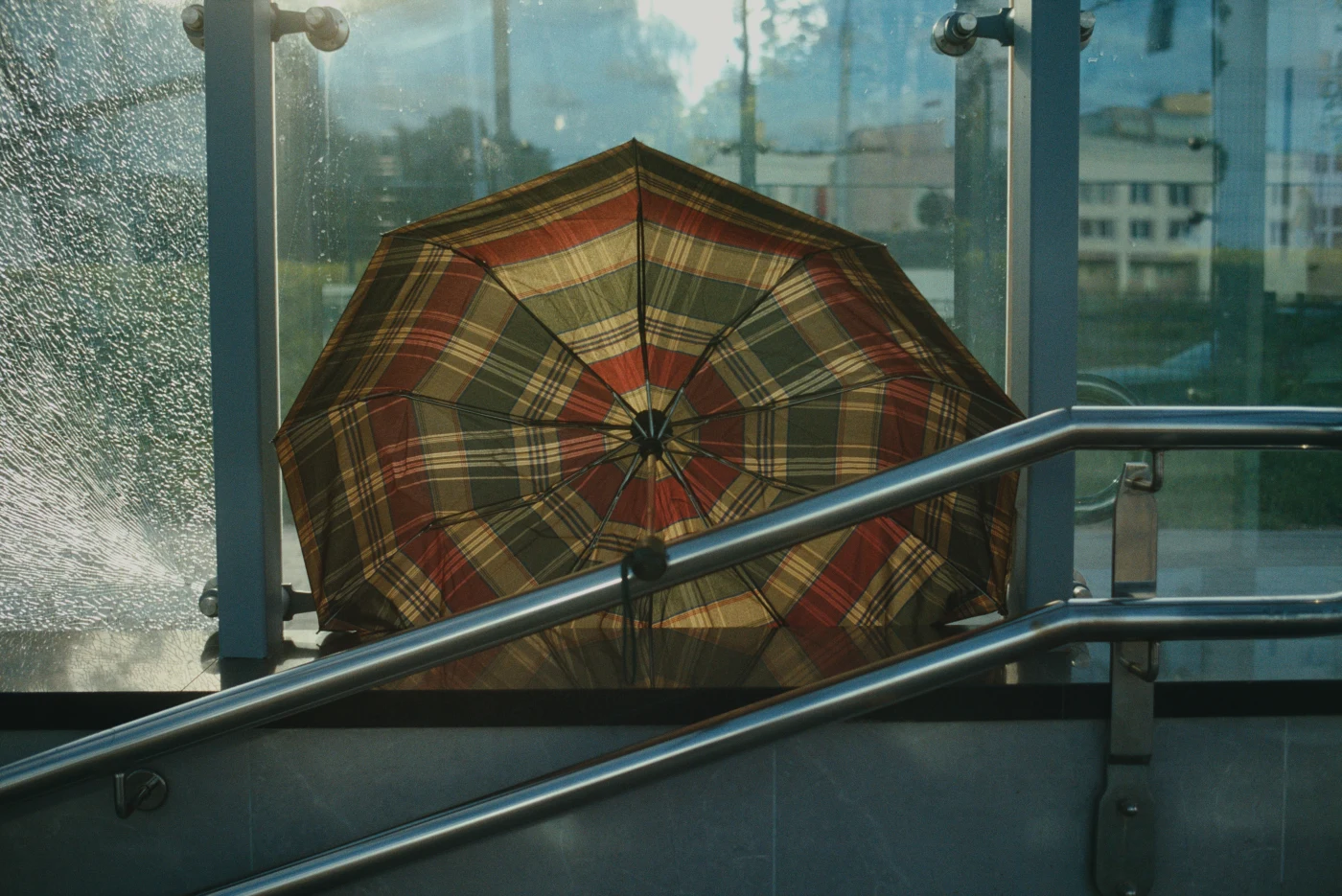An umbrella with a checkered pattern stands by a glass wall, with blurred buildings and sunlight in the background.