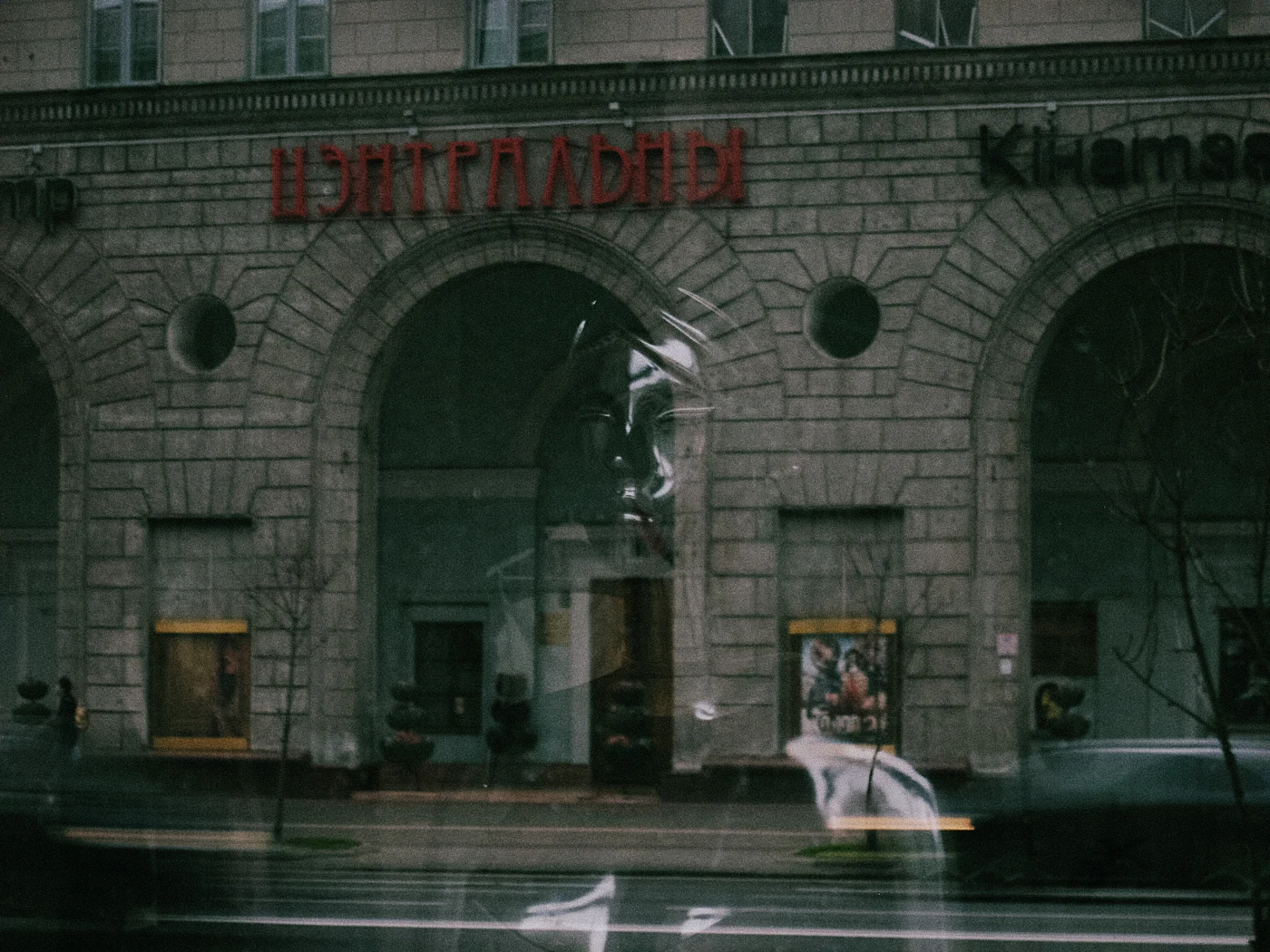 A person&rsquo;s reflection against a building with the sign “Centralny”. Shop windows and the street are out of focus.