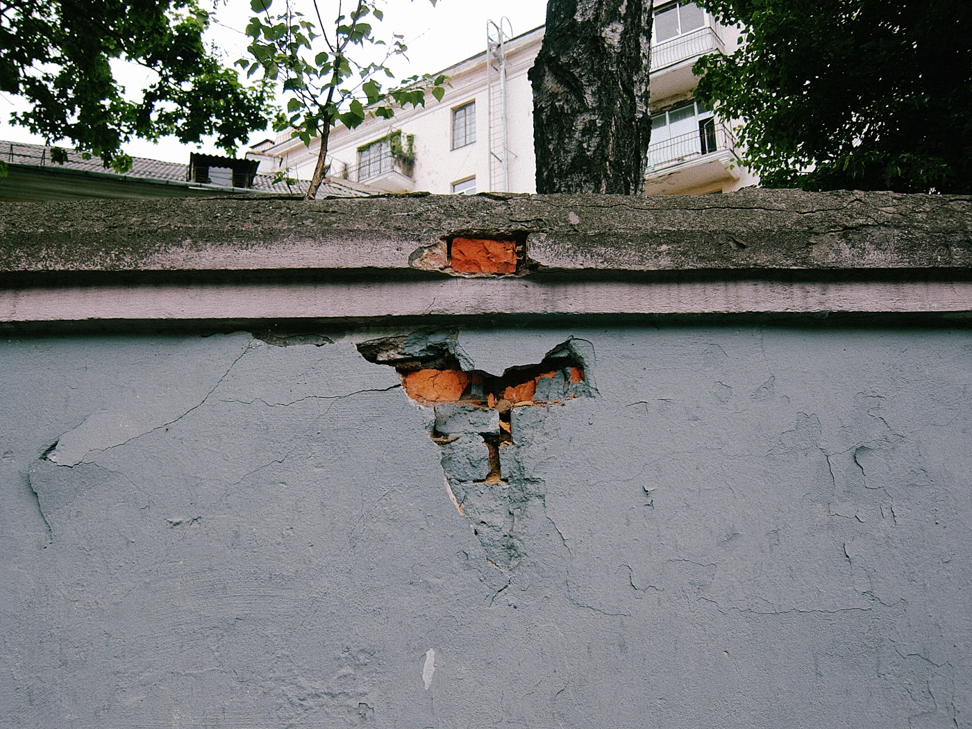 A crack in a wall with exposed brick, with green trees and buildings in the background.