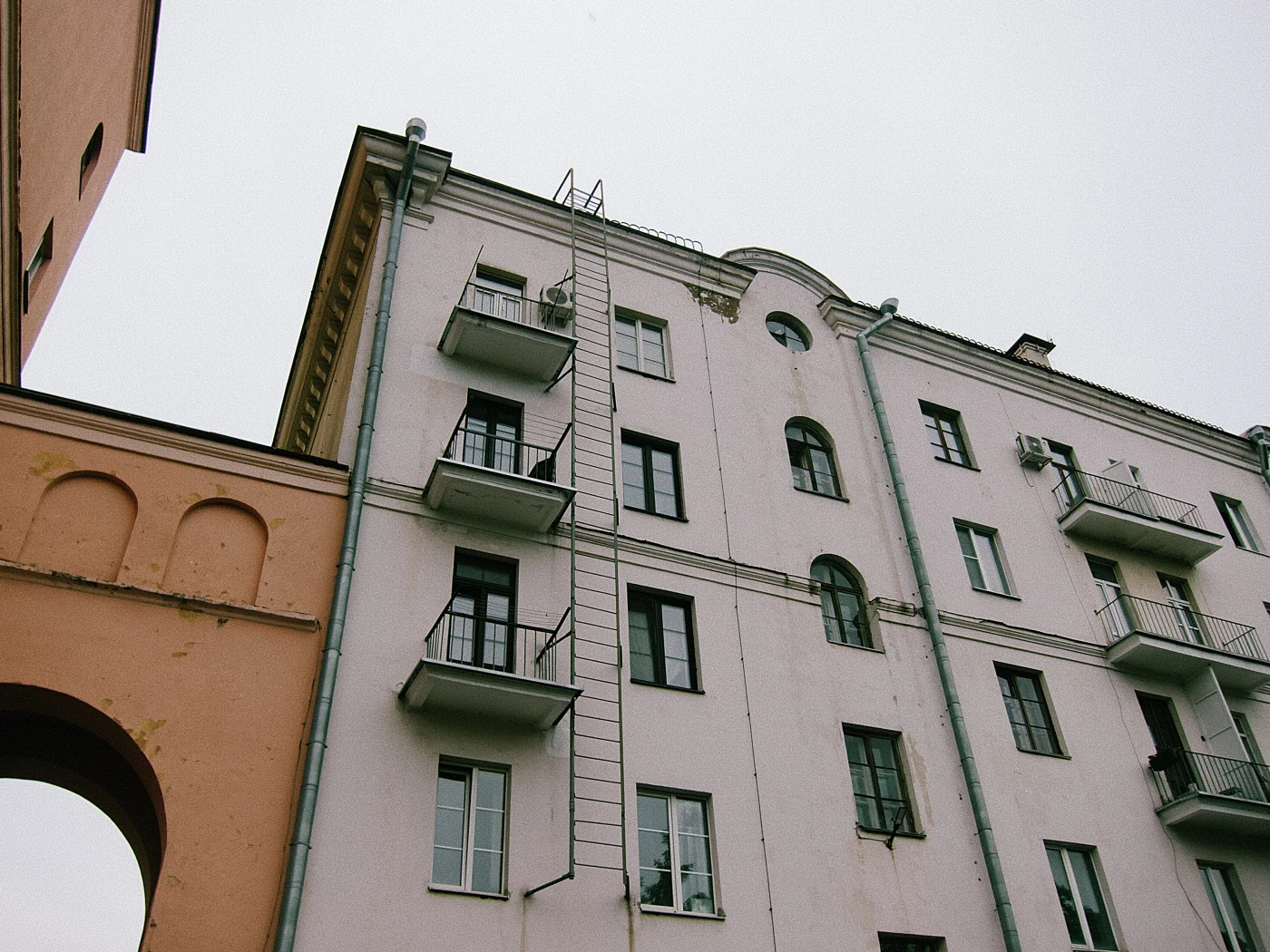 The facade of a building with balconies and a fire escape, gray in color, under a cloudy sky.