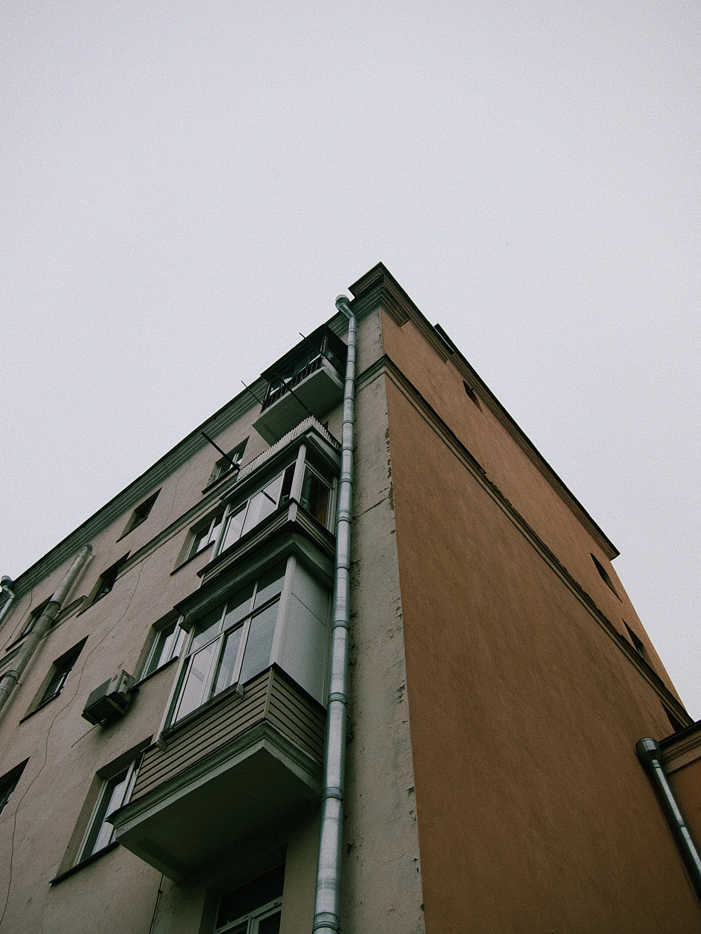 A high-rise building with balconies and a drainpipe, photographed from below against a gray sky.