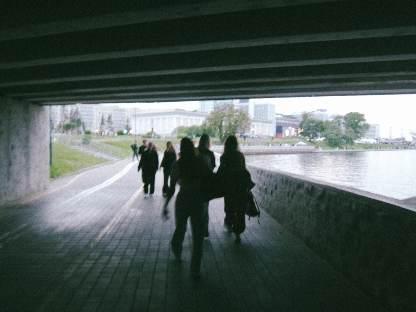 A group of people walking under a bridge, with an urban landscape and river visible in the background.