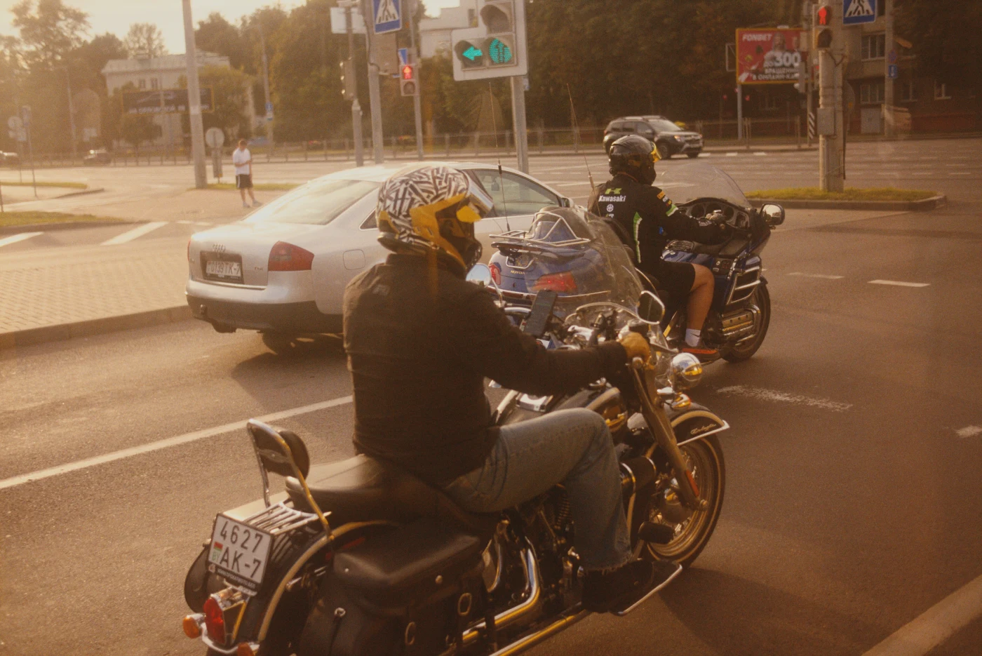 Motorcyclists at an intersection, one wearing a patterned helmet and another in sports gear. A car is visible in the background.
