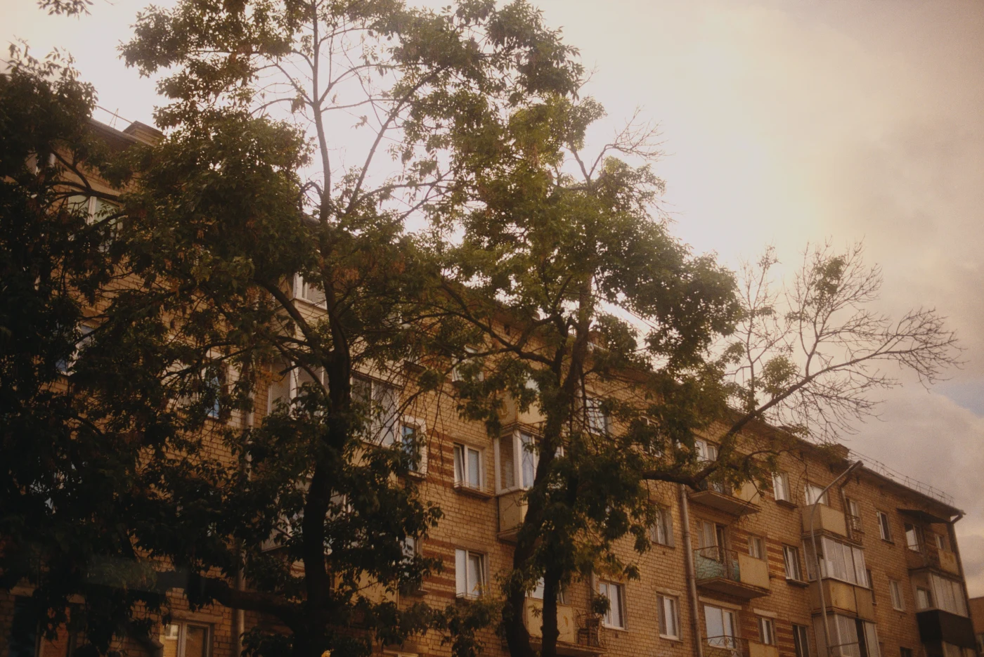 A residential building with balconies against a cloudy sky and trees. Sunlight illuminates the facade.