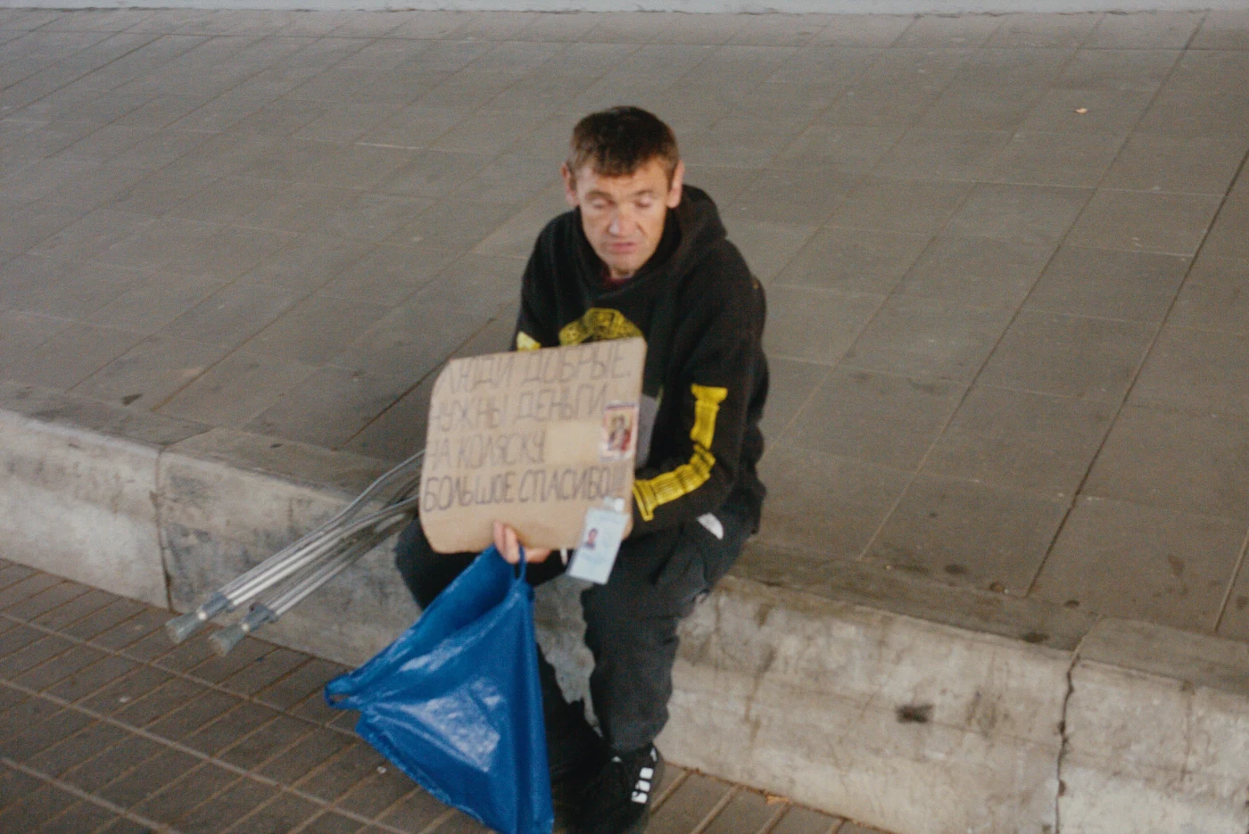 A person sits on the ground with a sign that reads: “Money needed for medication. Thank you very much.”