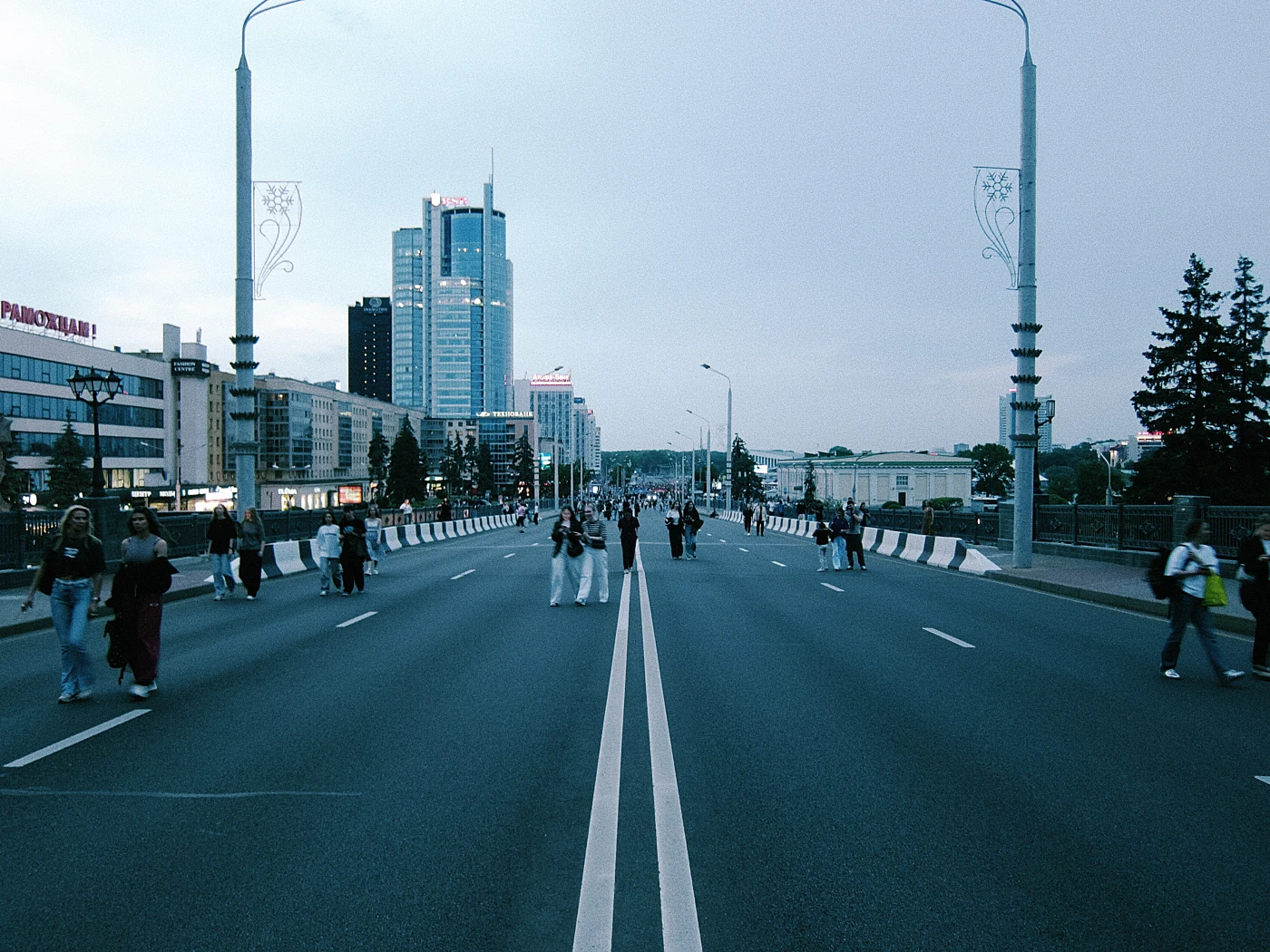 Pedestrians on a wide avenue, with buildings and city lights against the evening sky in the background.