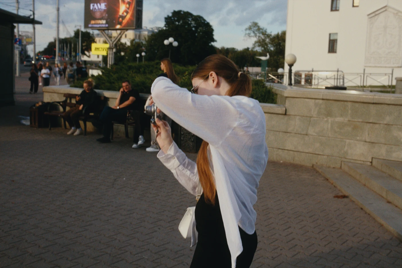 A woman takes a photo of herself on the street, with people and a billboard reading “FAME” in the background.