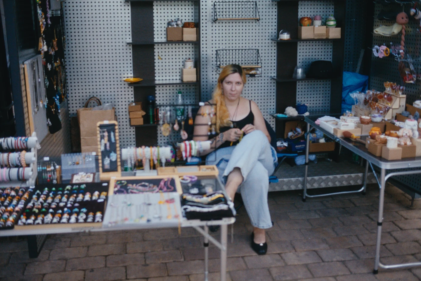 A woman with long hair sits behind a stall with handmade items and jewelry.