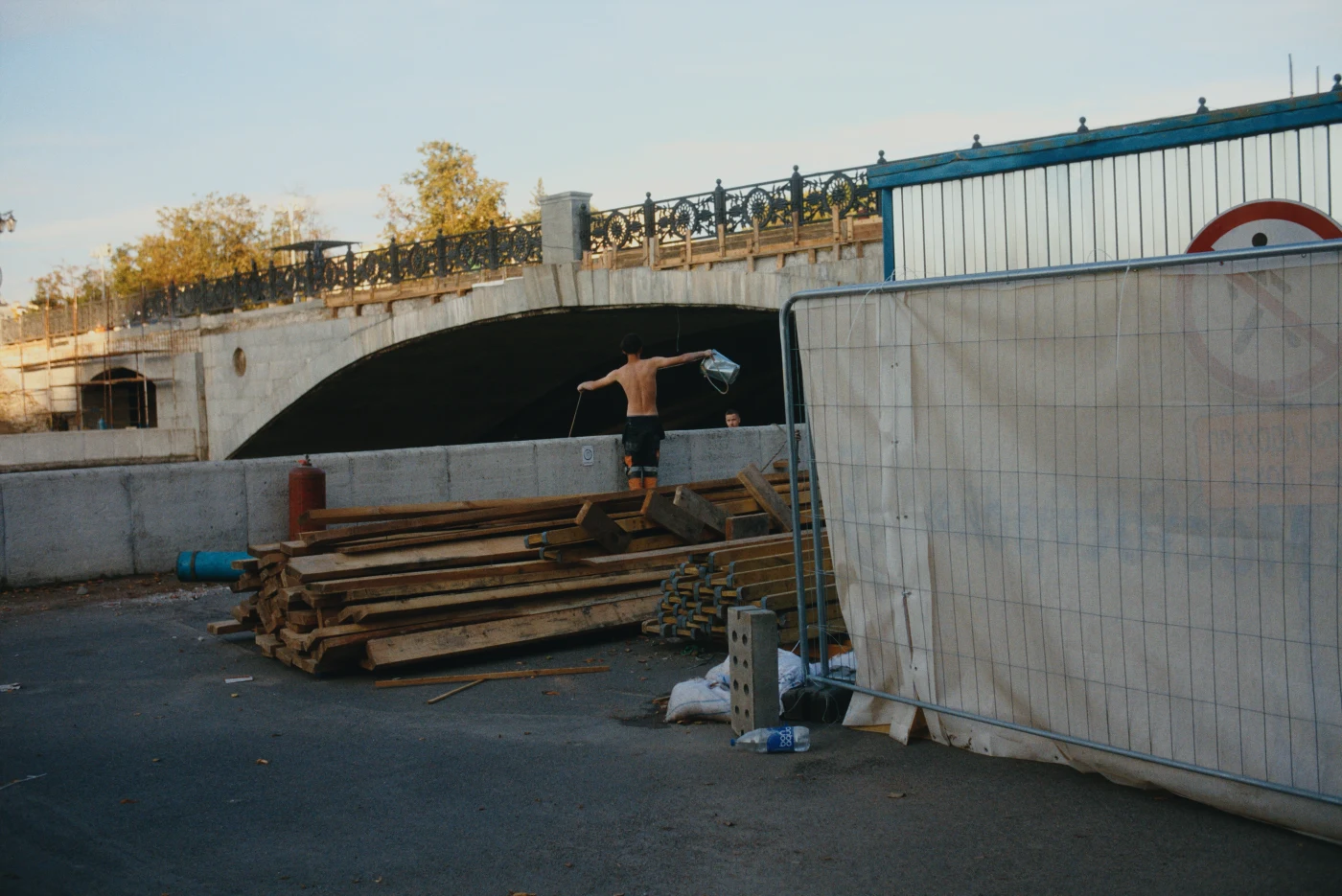 A shirtless man stands on construction materials near a bridge arch, with fencing and construction work nearby.