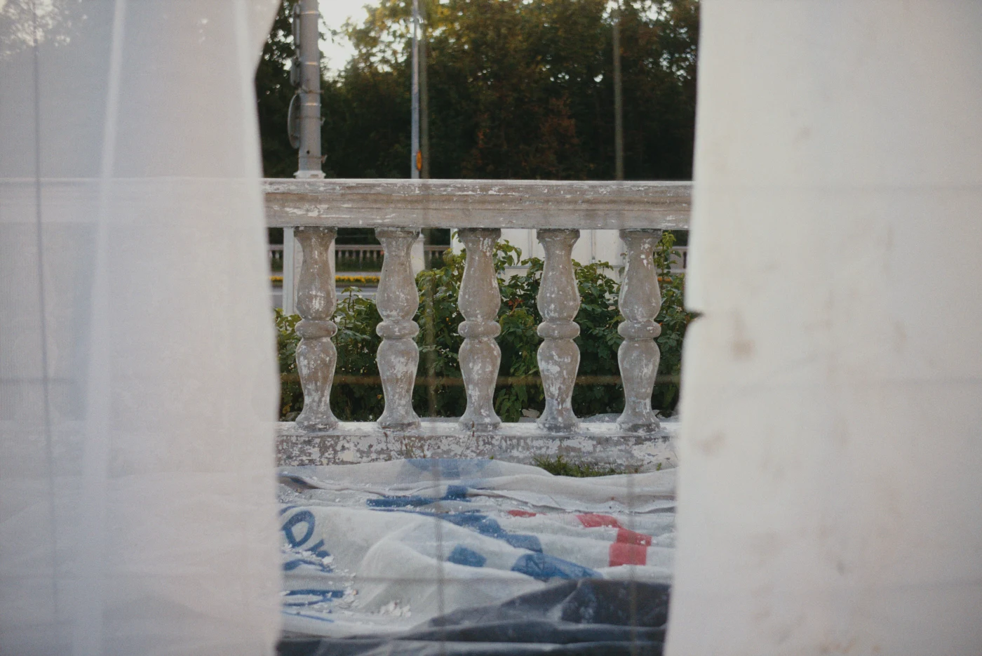 A curtain is partially open, revealing a balcony with columns and a floor covered in plastic sheeting. Greenery is visible in the background.