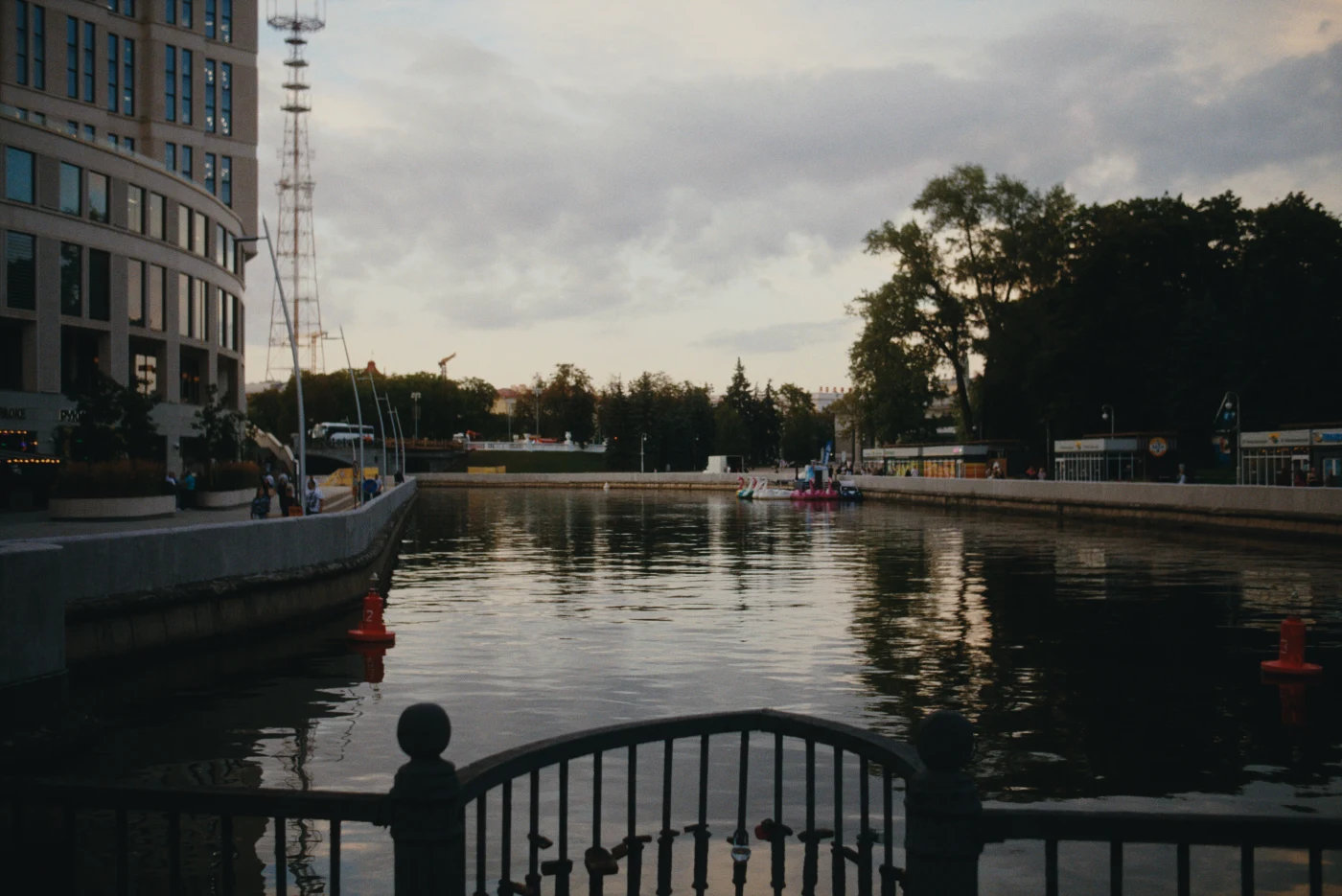 A calm river reflecting the clouds, with buildings and trees along the shore and boats visible in the distance.