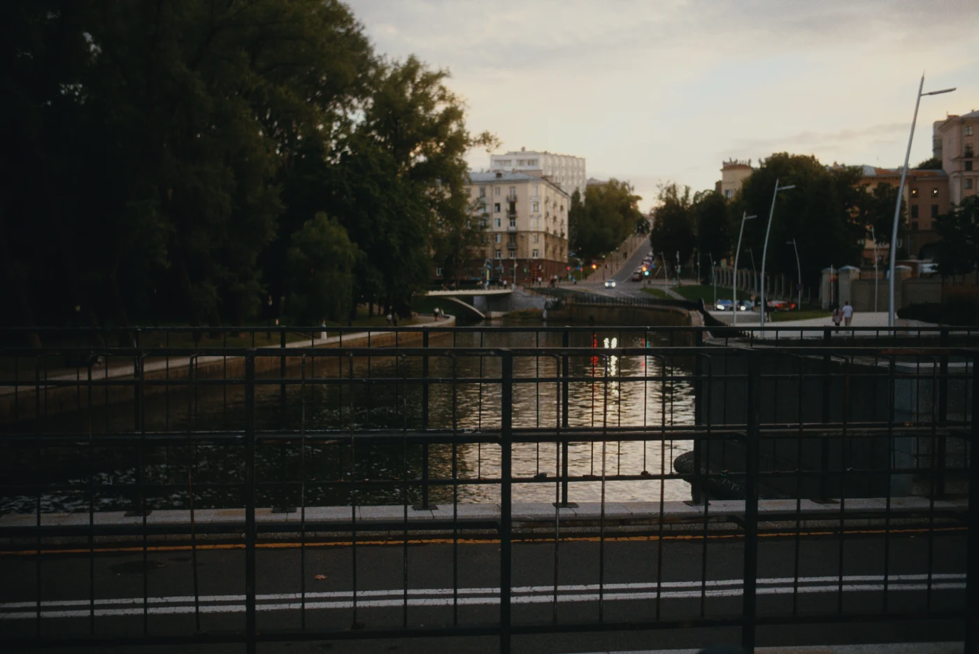 A view of a river and street with a railing, evening light, green trees, and buildings in the background.