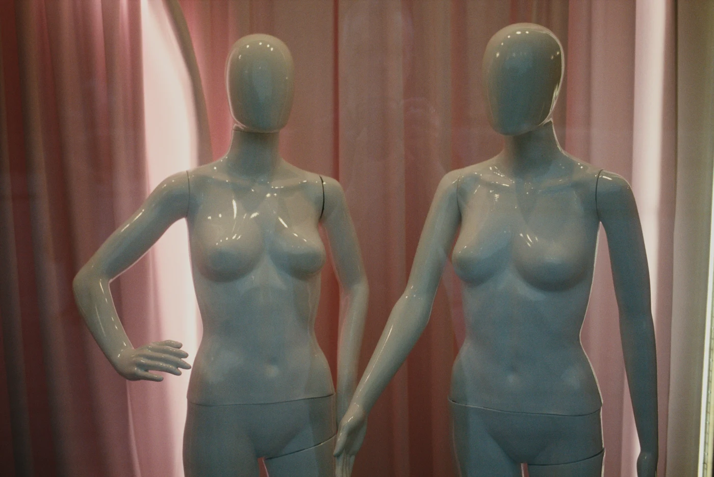 Two smooth-surfaced mannequins standing side by side against pink curtains.