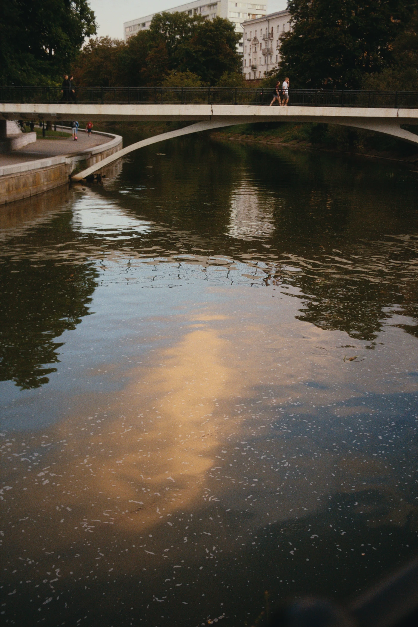 Clouds reflected in the water, with a bridge across the river and people on the bridge and along the shore. Green trees surround the scene.