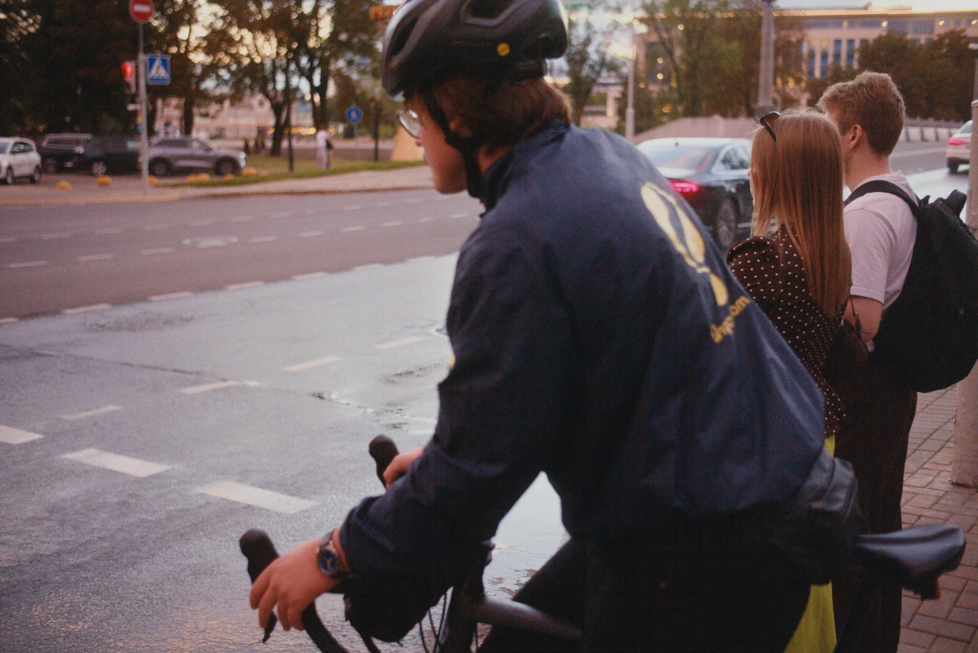 A cyclist wearing a helmet waits at a traffic light beside two people. It is raining and the pavement is wet.