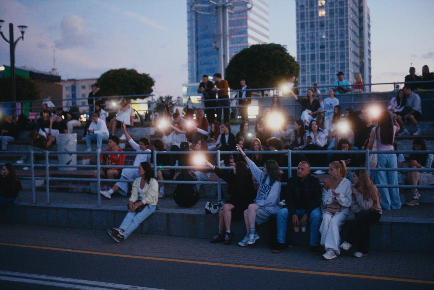 A group of people on benches, some filming with their phones, against an evening cityscape.