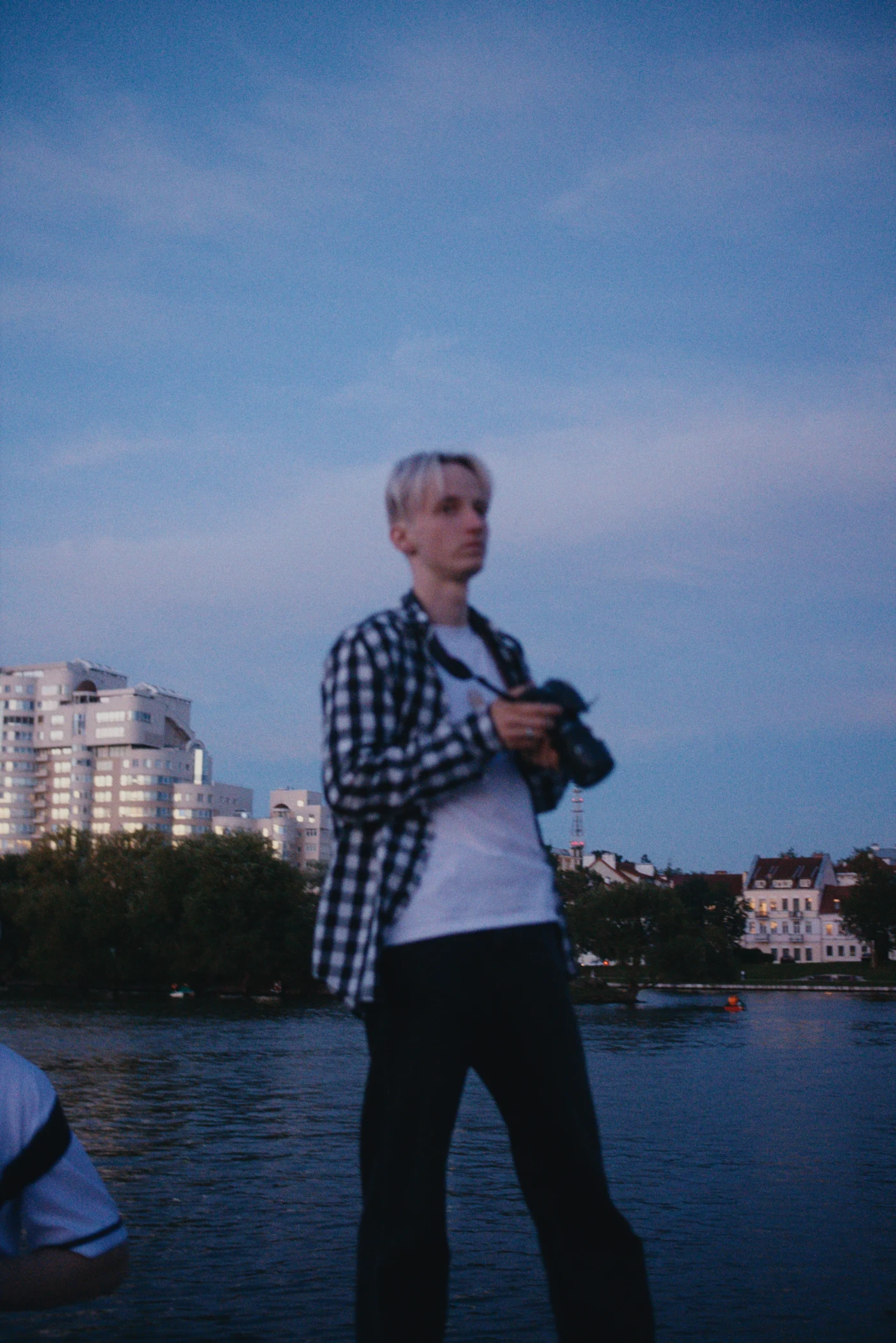 A man with a camera against a river and city buildings in the evening light.